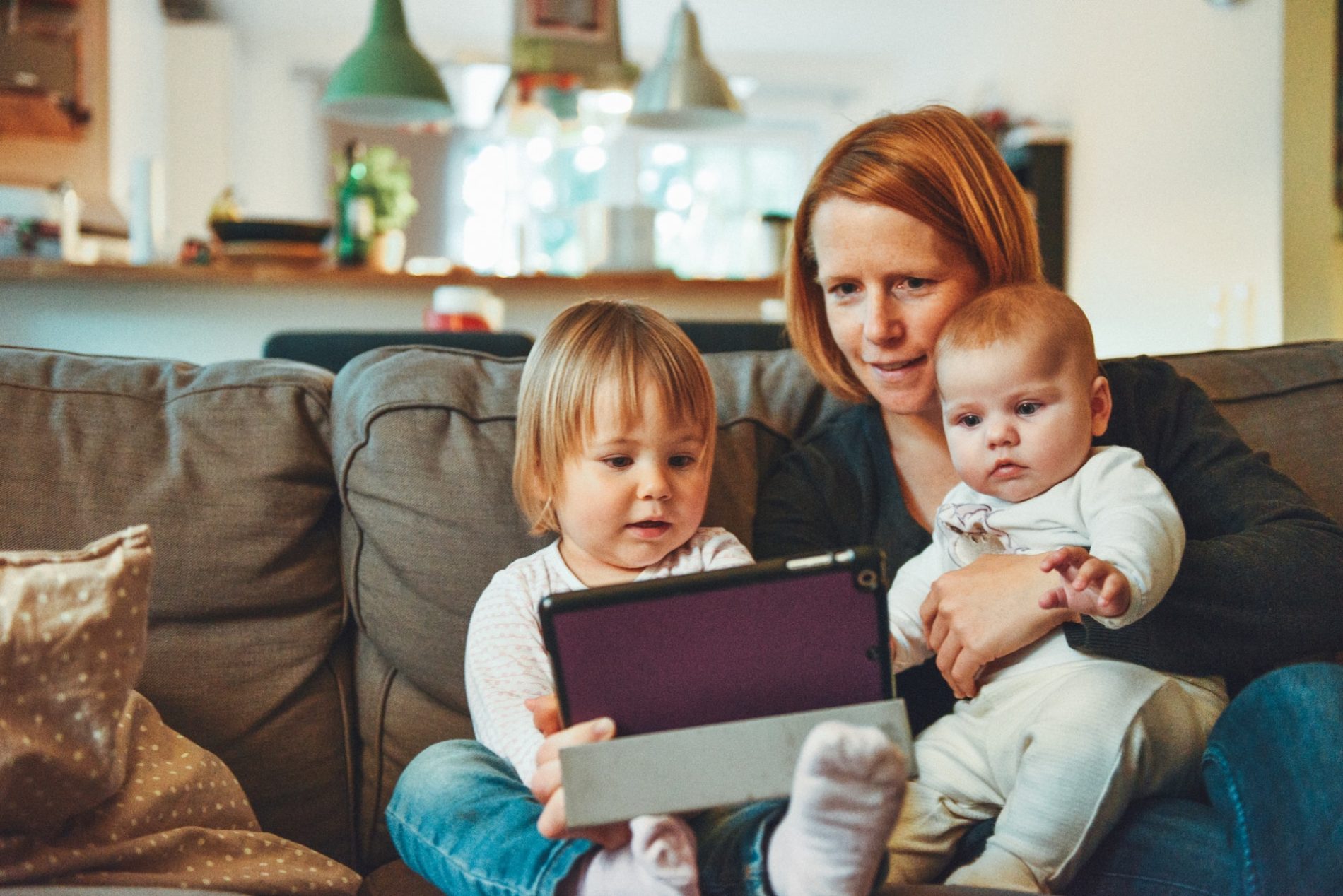 Mutter sitzt mit zwei Kleinkindern auf dem Sofa und zeigt ihnen etwas auf einem Tablet