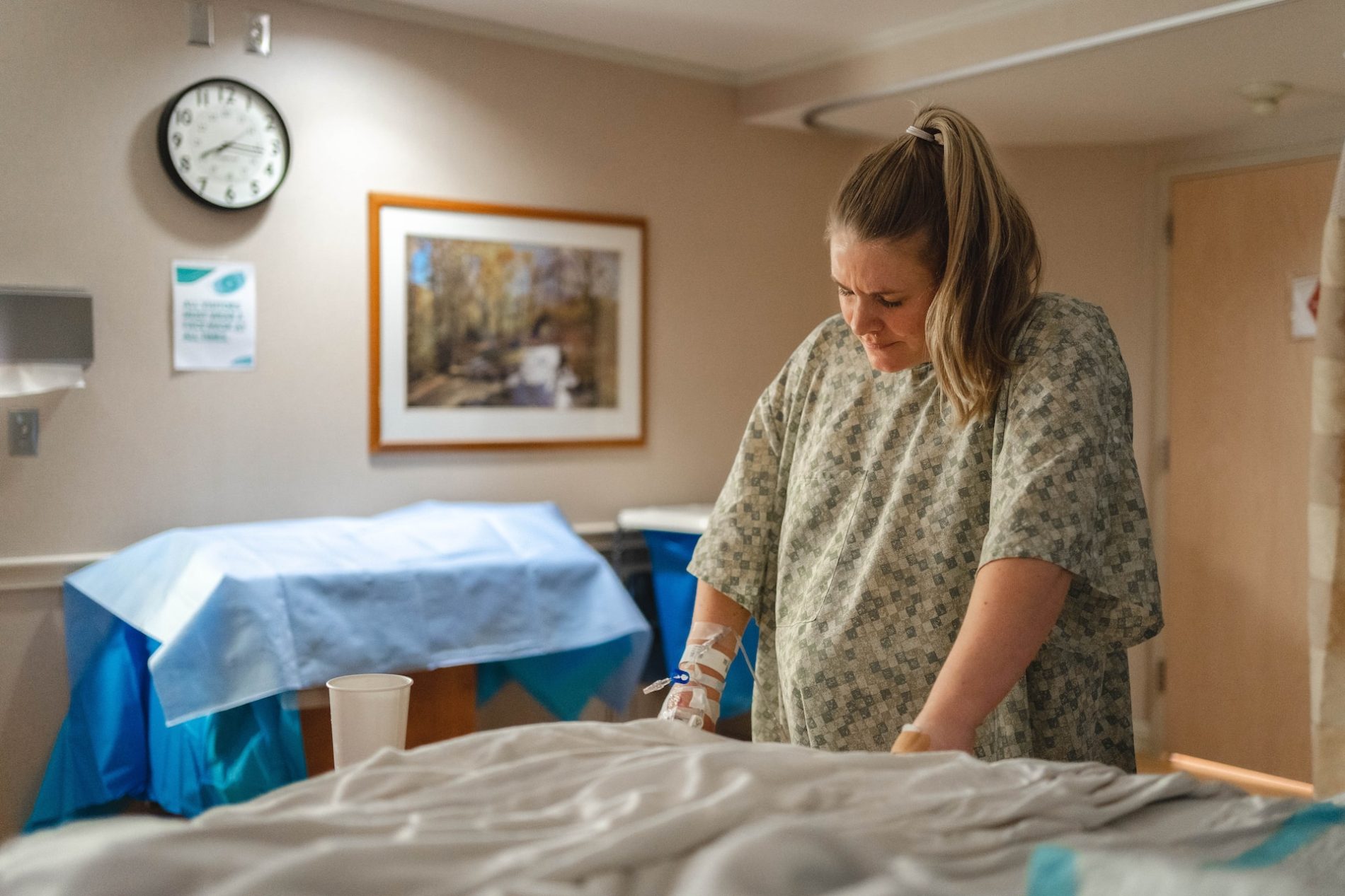 Eine hochschwangere Frau steht in einem grün gemusterten Nachthemd in ihrem Krankenhauszimmer vor ihrem Bett